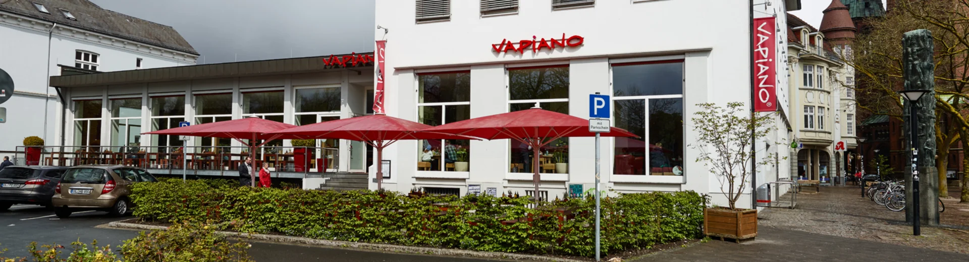 Außenansicht vom VAPIANO Oldenburg mit Bänken und roten Sonnenschirmen im Außenbereich.