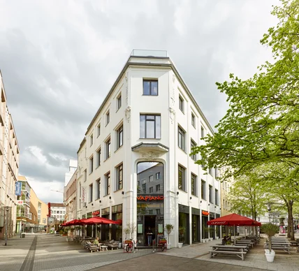Helles Eckgebäude mit klassischer Fassade; unten Restaurant mit Außenterrassen, roten Schirmen und Sitzbänken an Fußgängerzone