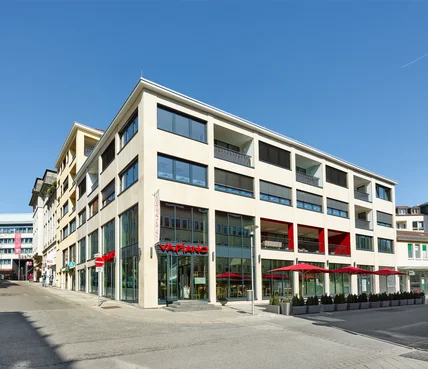 Modernes Eckgebäude mit heller Fassade und großen Fenstern; unten Restaurantterrasse mit roten Schirmen an ruhiger Straße