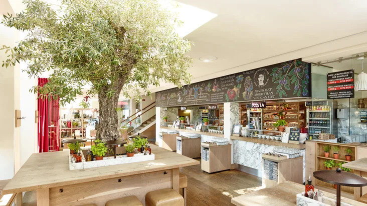 Innenaufnahme vom VAPIANO Leipzig mit Tischen, einem Olivenbaum und der Theke zu Bestellen im Hintergrund.