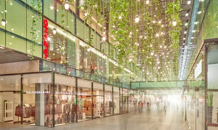 Modernes Einkaufszentrum mit Glasfassaden; hohe Decke mit vielen Lichtern und hängenden Pflanzen, Geschäfte entlang des Gangs