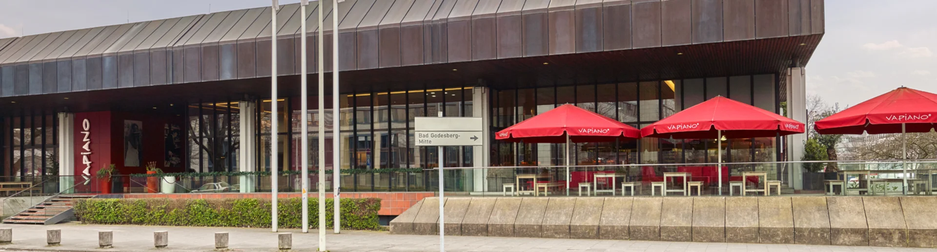 Außenansicht vom VAPIANO Bonn mit Bänken, Tischen und roten Sonnenschirmen im Außenbereich.