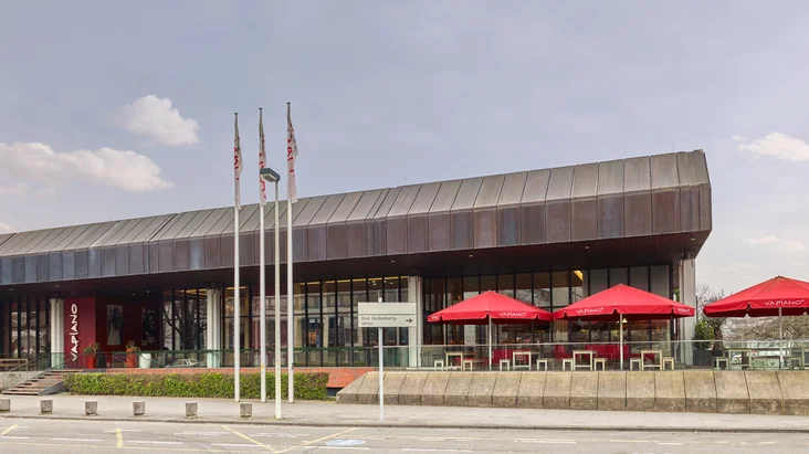 Außenansicht vom VAPIANO Bonn mit Bänken, Tischen und roten Sonnenschirmen im Außenbereich.