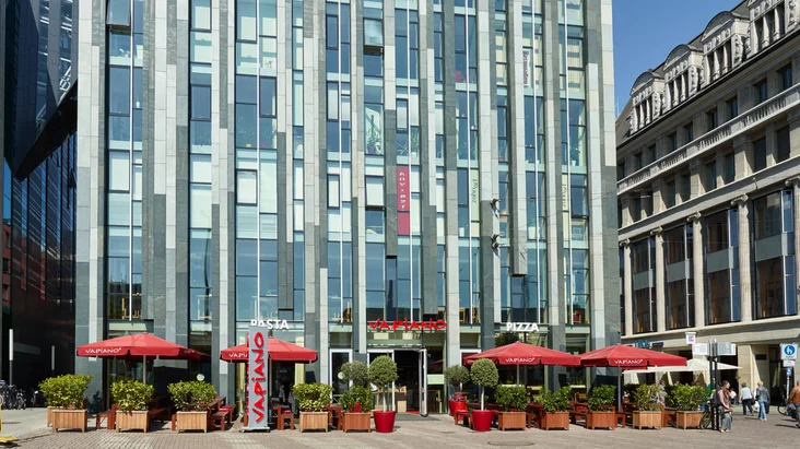 Außenansicht vom VAPIANO Leipzig im Erdgeschoss eines modernen Gebäudes mit Bänken und Sonnenschirmen.