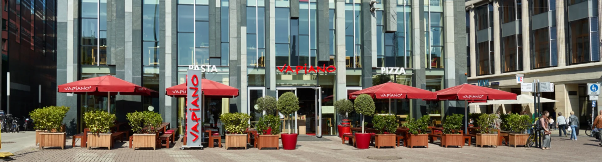 Außenansicht vom VAPIANO Leipzig im Erdgeschoss eines modernen Gebäudes mit Bänken und Sonnenschirmen.