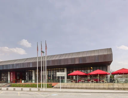 Flaches modernes Gebäude mit Glasfront und Metalldach; davor Terrasse mit roten Schirmen und Fahnenmasten an Straße