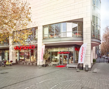 Modernes Gebäude mit heller Steinfassade; Restauranteingang mit Glasfront, Fahnen, Sitzbänken und breitem Gehweg davor