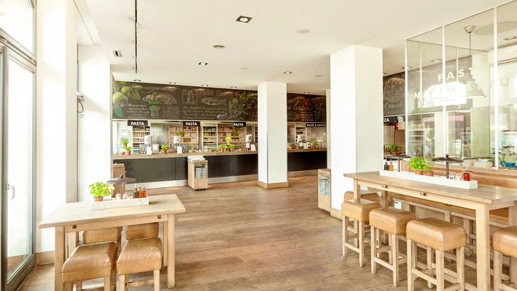 Innenaufnahme vom VAPIANO Mannheim mit Tischen, Hockern und der Theke zum Bestellen im Hintergrund.
