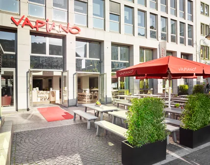 Restaurantfassade mit großen Fenstern; Außenterrasse mit Holzbänken, Pflanzenkübeln und roten Sonnenschirmen