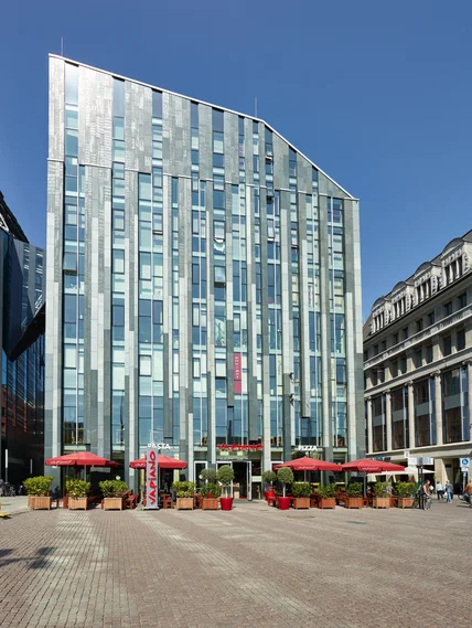 Hohes modernes Glasgebäude mit schräger Dachkante; großer Platz mit Pflanzenkübeln und Restaurantterrasse mit roten Schirmen