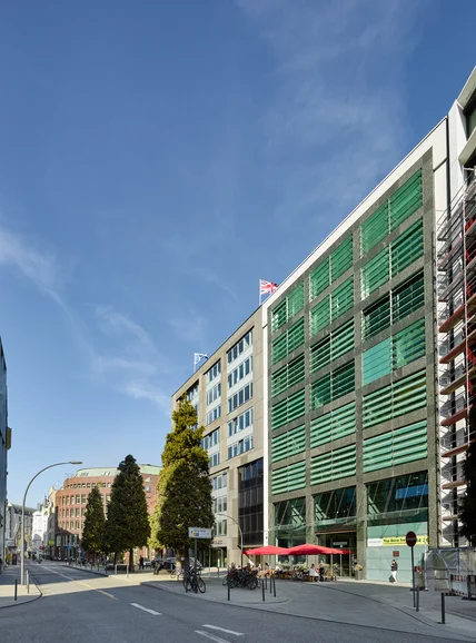 Modernes Bürogebäude mit grüner Lamellenfassade; Straßenecke mit Bäumen, Fahrrädern und Caféterrasse mit roten Schirmen