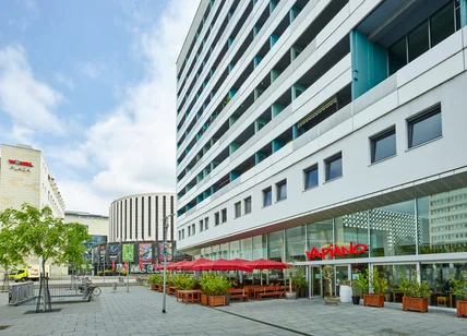 Hohes modernes Gebäude mit vielen Fenstern; Erdgeschoss Restaurant mit Terrasse, roten Schirmen und Pflanzkästen am Platz