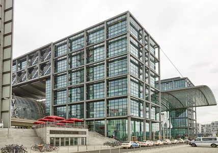 Modernes Glasgebäude mit Stahlrahmen, großem Vordach; Treppenaufgang, Fahrräder, Taxis und rote Sonnenschirme davor