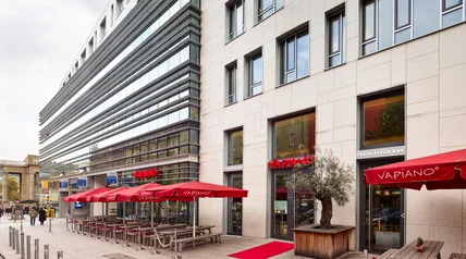 Modernes Gebäude mit Stein- und Glasfassade; Restaurantterrasse mit roten Schirmen, Holztischen und Pflanzkübel an Straße