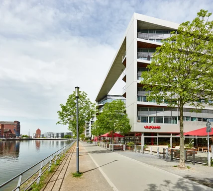 Modernes Gebäude am Fluss mit breitem Dachüberstand; Uferpromenade, Bäume und Restaurantterrasse mit roten Schirmen