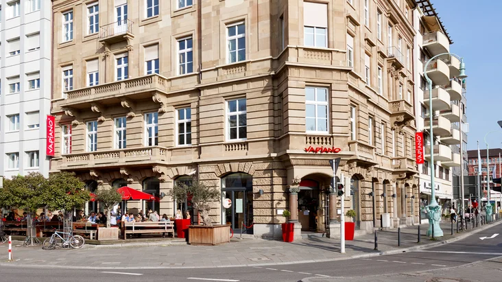 Außenansicht vom VAPIANO Mannheim im Erdgeschoss eines historischen Gebäudes mit Bänken und Sonnenschirmen.