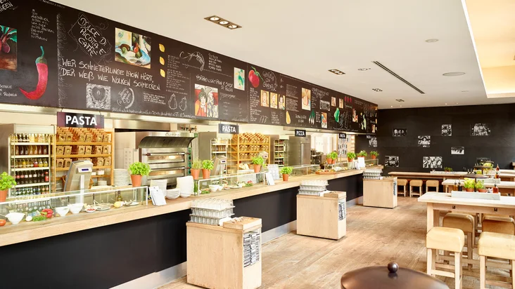 Innenaufnahme vom VAPIANO Köln Deutz mit Tischen und Stühlen und der Theke zum Bestellen im Hintergrund.