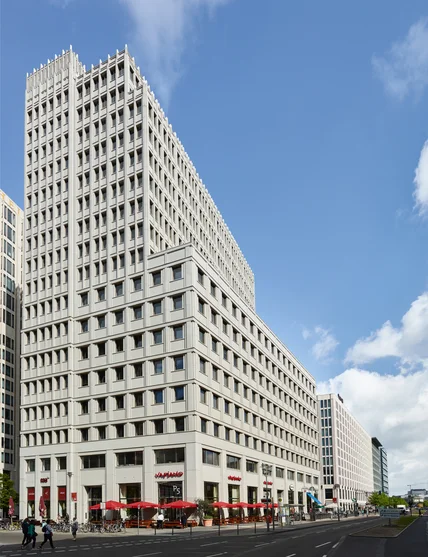 Hohes modernes Bürogebäude mit gestufter Fassade, vielen Fenstern; unten Cafés mit roten Schirmen an breiter Straße