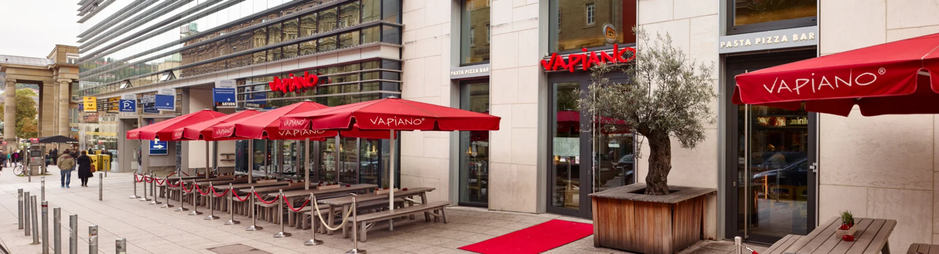 Außenansicht vom VAPIANO Stuttgart mit Bänken, Tischen und roten Sonnenschirmen in der Fußgängerzone.