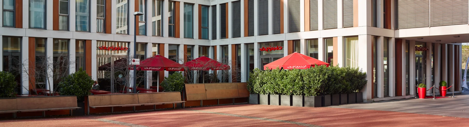 Außenansicht vom VAPIANO Köln Deutz im Erdgeschoss eines mehrstöckigen Gebäudes mit Bänken und roten Sonnenschirmen.