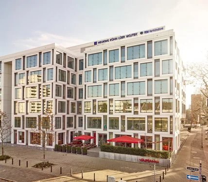 Modernes Bürogebäude mit heller Rasterfassade und großen Fenstern; unten Restaurantterrasse mit roten Schirmen an ruhiger Straße