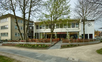 Flaches modernes Gebäude mit heller Fassade; große Außenterrasse mit Holztischen, Bäumen und breitem Gehweg davor