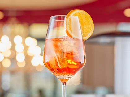 Ein Aperol Sprizz mit Eiswürfel und einer Orangenscheibe steht auf einer Bar, stilvoll in Szene gesetzt vor einem warm beleuchteten Hintergrund.