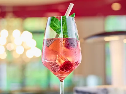 Ein roséfarbener, spritziger Drink mit Beeren, Minze und Strohhalm steht auf einer Bar, stilvoll in Szene gesetzt vor einem warm beleuchteten Hintergrund.